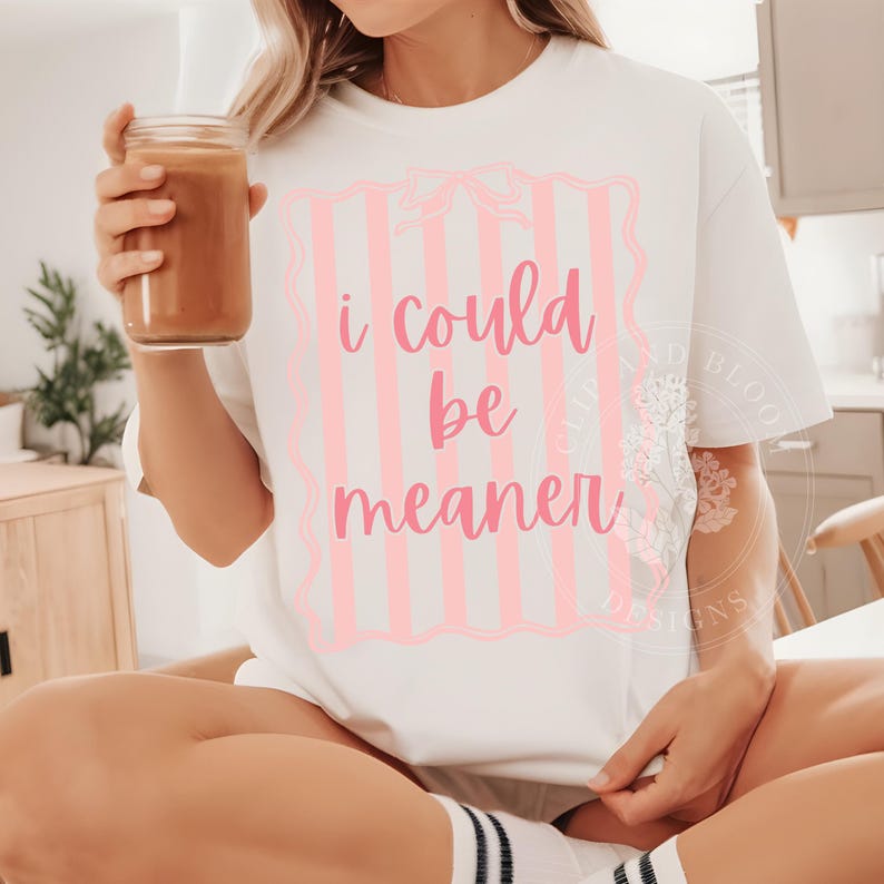May include: White t-shirt with the text "I could be meaner" in pink script. The text is inside a pink striped frame with a bow at the top. The person is holding a glass of brown liquid.