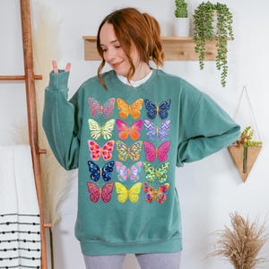 May include: Teal-colored sweatshirt featuring a vibrant array of colorful butterfly designs. The butterflies are in various colors and patterns, including pink, yellow, blue, and orange. The sweatshirt has a crew neck and long sleeves, offering a casual and comfortable style.