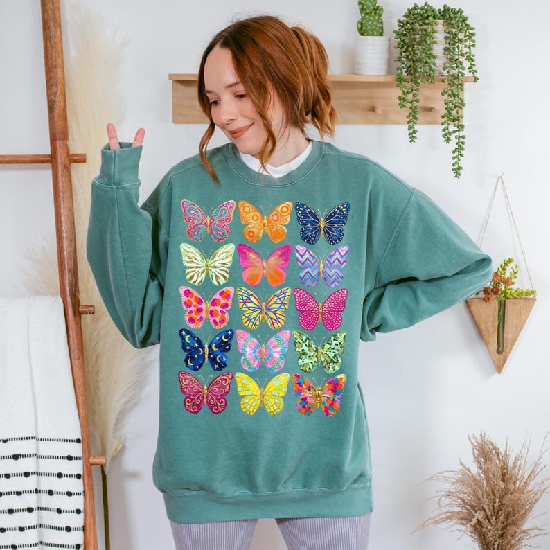 May include: Teal-colored sweatshirt featuring a vibrant array of colorful butterfly designs. The butterflies are in various colors and patterns, including pink, yellow, blue, and orange. The sweatshirt has a crew neck and long sleeves, offering a casual and comfortable style.