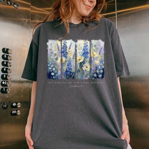 Puede incluir: Camiseta gris oscuro con un dise&ntilde;o floral con flores azules, amarillas y blancas. El texto "HIS MERCIES ARE NEW EVERY MORNING" est&aacute; impreso debajo. La camiseta est&aacute; hecha de un material suave.
