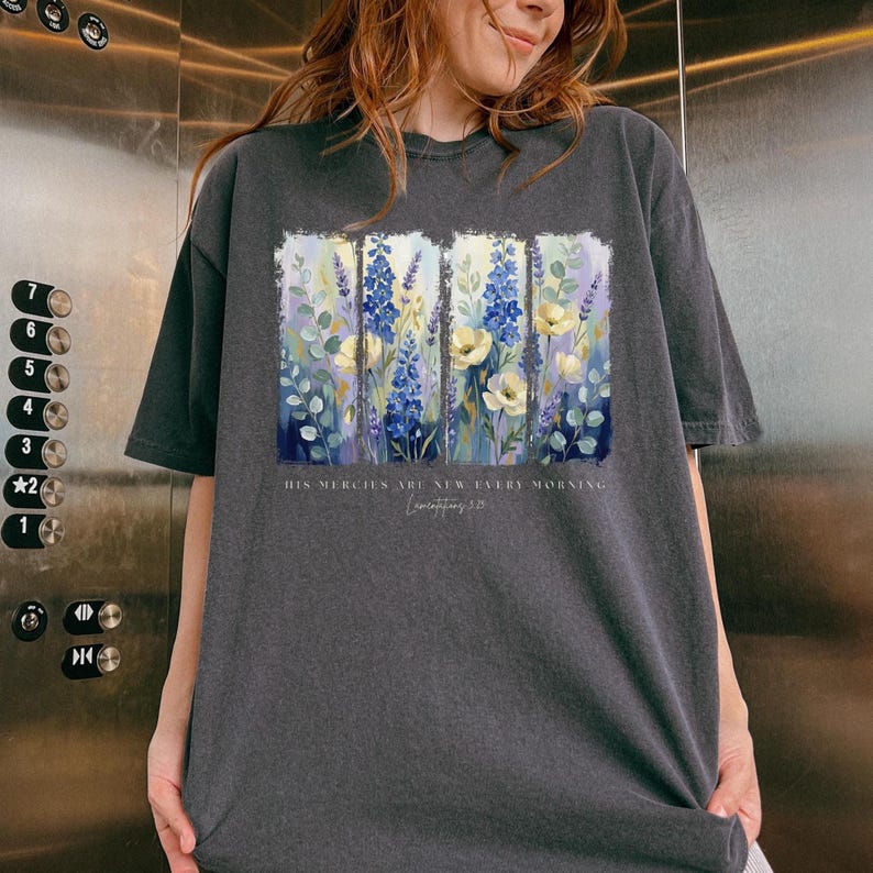 Puede incluir: Camiseta gris oscuro con un dise&ntilde;o floral con flores azules, amarillas y blancas. El texto "HIS MERCIES ARE NEW EVERY MORNING" est&aacute; impreso debajo. La camiseta est&aacute; hecha de un material suave.