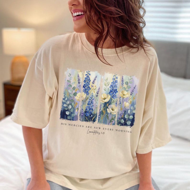 Puede incluir: Camiseta color crema con un dise&ntilde;o floral con flores azules y blancas. La camiseta tiene mangas cortas y el texto "HIS MERCIES ARE NEW EVERY MORNING" y "Lamentations 3:23".