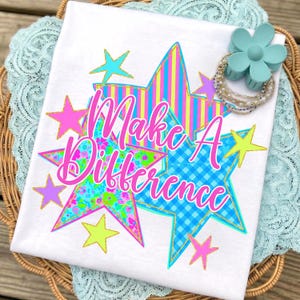 May include: A white t-shirt featuring a graphic of overlapping stars in yellow, blue, and pink patterns. The text "Make A Difference" is printed in bright pink script across the center. A light blue flower-shaped hair clip and beaded bracelets sit on top.