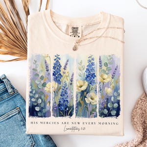 Puede incluir: Camiseta color crema con un dise&ntilde;o floral con flores azules y blancas. La camiseta tiene el texto "HIS MERCIES ARE NEW EVERY MORNING" y "Lamentations 3:23". Un collar plateado est&aacute; drapeado sobre la camiseta.