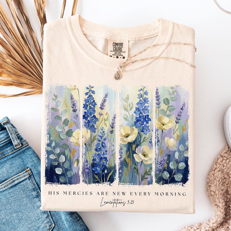 Puede incluir: Camiseta color crema con un dise&ntilde;o floral con flores azules y blancas. La camiseta tiene el texto "HIS MERCIES ARE NEW EVERY MORNING" y "Lamentations 3:23". Un collar plateado est&aacute; drapeado sobre la camiseta.