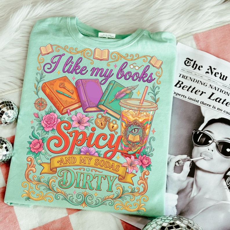 May include: A light green t-shirt with a colorful graphic design. The design includes the text "I like my books Spicy and my sodas Dirty" with illustrations of books, flowers, and a drink. The shirt is folded on a white surface.