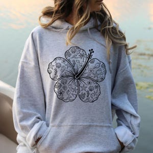 May include: A gray hoodie featuring a large black hibiscus flower design. The flower's petals are filled with various sea-themed illustrations, including seashells, starfish, and seahorses. The hoodie has a drawstring hood and a front pocket.