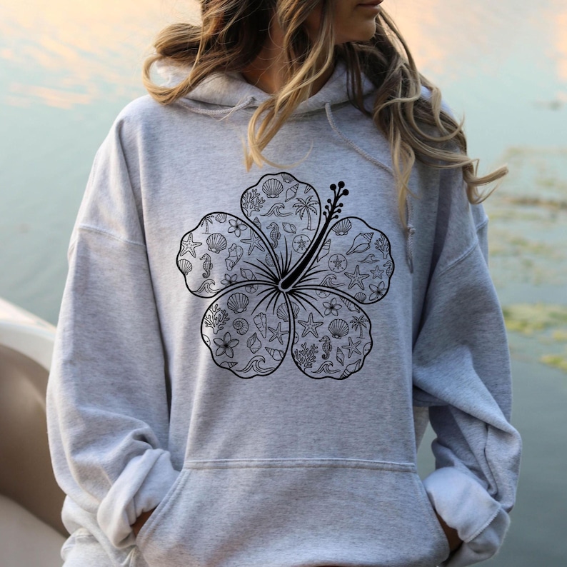 May include: A gray hoodie featuring a large black hibiscus flower design. The flower's petals are filled with various sea-themed illustrations, including seashells, starfish, and seahorses. The hoodie has a drawstring hood and a front pocket.