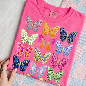 May include: A pink t-shirt featuring a collection of colorful butterfly illustrations. The butterflies are in various designs and colors, including blue, orange, pink, and green. The shirt has a crew neck and a tag that says "Comfort Colors."