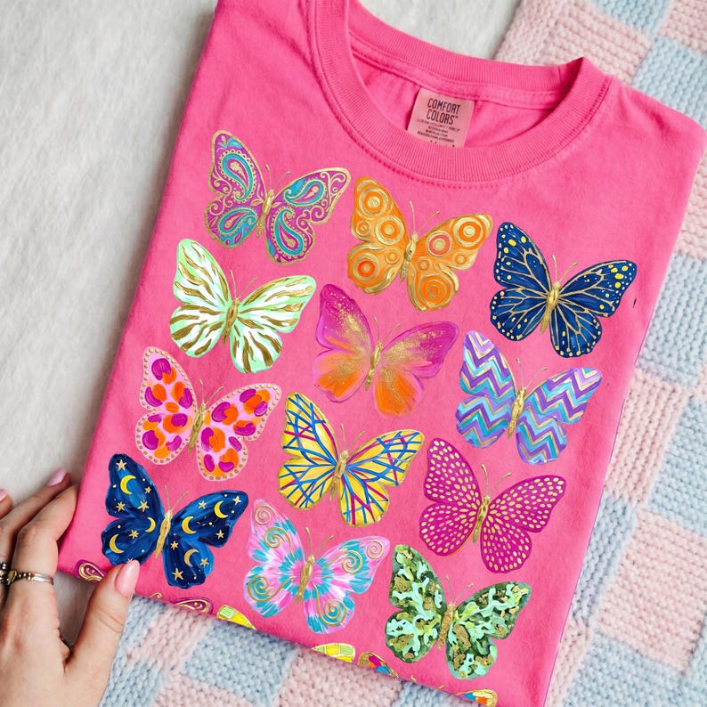 May include: A pink t-shirt featuring a collection of colorful butterfly illustrations. The butterflies are in various designs and colors, including blue, orange, pink, and green. The shirt has a crew neck and a tag that says "Comfort Colors."
