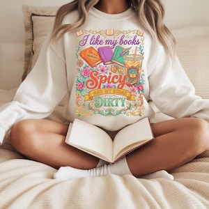 May include: Cream-colored sweatshirt with a colorful graphic that reads "I like my books spicy and my sodas dirty." The graphic includes illustrations of books, flowers, and a drink. The person is sitting with an open book.