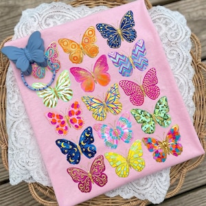 May include: Pink fabric with a collection of colorful butterfly designs. The butterflies feature various patterns and colors, including blue, orange, pink, and yellow. A blue butterfly-shaped pacifier clip with a beaded chain is attached to the fabric.
