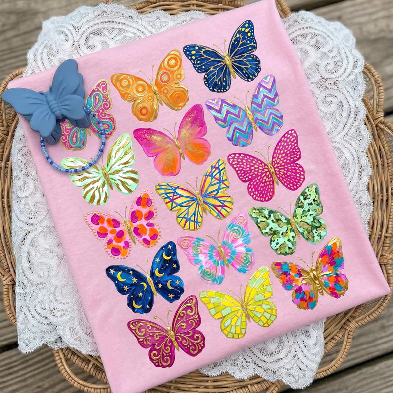May include: Pink fabric with a collection of colorful butterfly designs. The butterflies feature various patterns and colors, including blue, orange, pink, and yellow. A blue butterfly-shaped pacifier clip with a beaded chain is attached to the fabric.