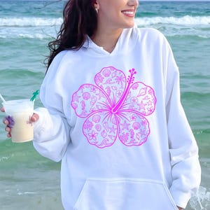 May include: White hoodie featuring a large, bright pink hibiscus flower design. The flower is filled with seashell and ocean-themed illustrations. The hoodie has a front pocket and a drawstring hood, perfect for casual wear.