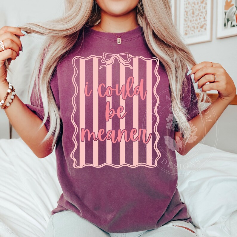 May include: A purple t-shirt with the text "I could be meaner" in a pink, striped frame. The shirt has short sleeves and a relaxed fit. The text is in a cursive font.