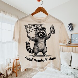 Pu&ograve; includere: T-shirt color crema con grafica di procione. Il procione tiene in mano una bandiera con la scritta "Trash Panda" e una bibita. Il testo "Feral Baseball Mom" &egrave; sotto il procione.