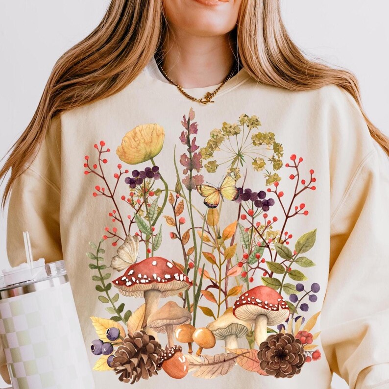 Peut inclure: Sweat-shirt cr&egrave;me avec un motif floral et champignon color&eacute;. Le graphisme comprend des champignons &agrave; chapeau rouge, des papillons jaunes et diverses fleurs et feuillages dans des tons de rouge, violet et vert. Un v&ecirc;tement confortable sur le th&egrave;me de la nature.