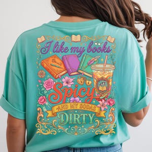 May include: Teal t-shirt with a colorful graphic that reads "I like my books Spicy and my sodas Dirty." The design includes books, flowers, and a drink with a straw. The shirt has short sleeves.