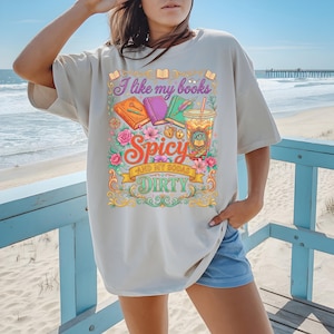 May include: Beige t-shirt with a colorful graphic that reads "I like my books spicy and my sodas dirty." The design includes illustrations of books, a drink, and floral elements. The shirt is a relaxed fit.