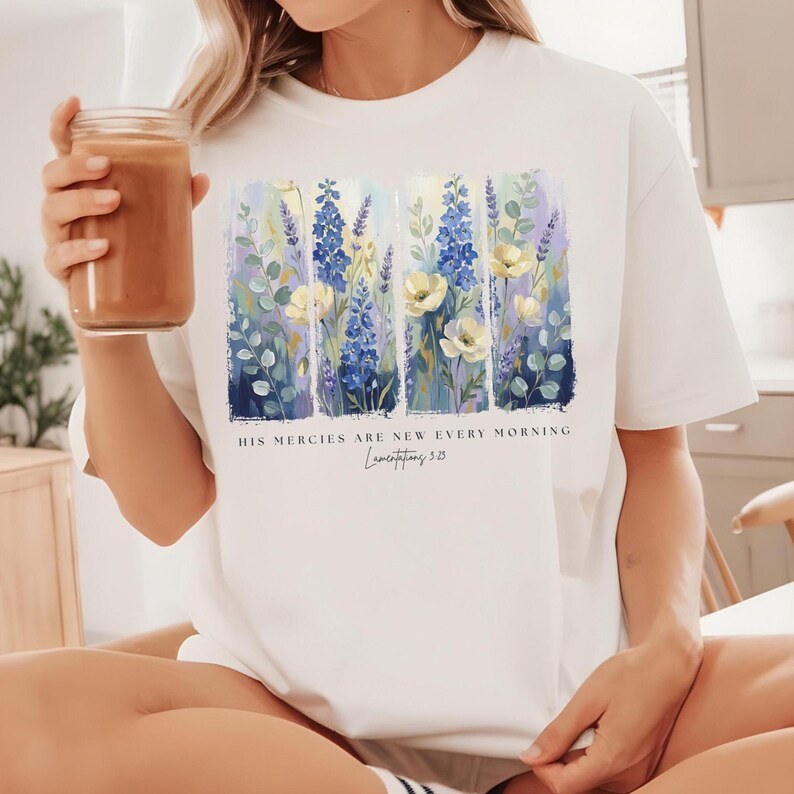 Puede incluir: Camiseta blanca con un dise&ntilde;o floral con flores azules, amarillas y moradas. La camiseta tiene el texto "HIS MERCIES ARE NEW EVERY MORNING" y "Lamentations 3:23". Una prenda casual para el d&iacute;a a d&iacute;a.