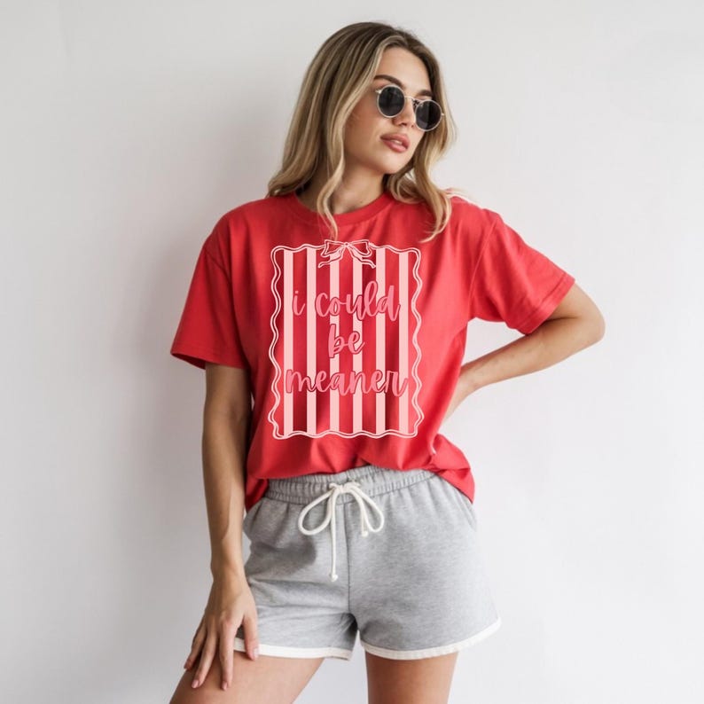 May include: A red t-shirt with the text "I could be meaner" in a pink and white striped design. The person is also wearing gray shorts with a white trim.