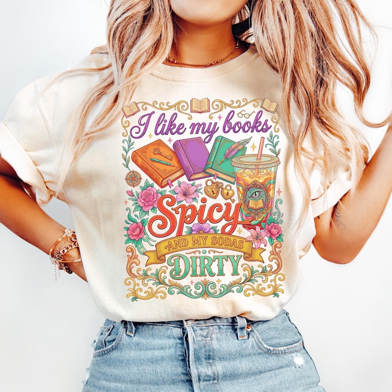 May include: Cream-colored t-shirt with a colorful graphic design. The design features the text "I like my books Spicy and my sodas Dirty" with illustrations of books, flowers, and a drink. The shirt has short sleeves and a crew neck.