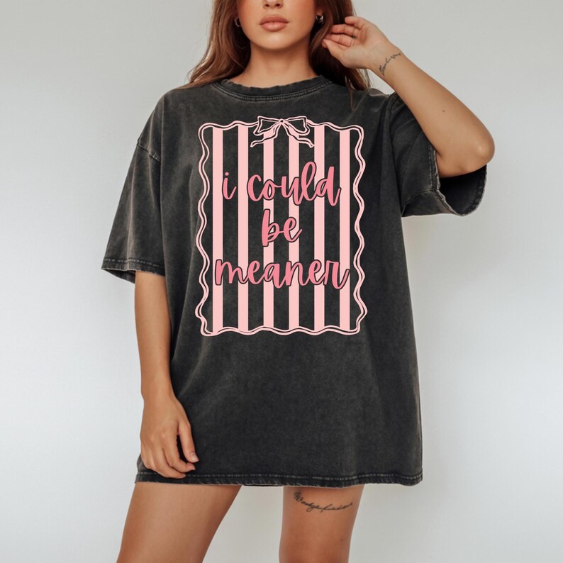 May include: Dark gray oversized t-shirt with a pink and white striped design. The design features the text "i could be meaner" in a pink script font, framed by a decorative border and a bow.