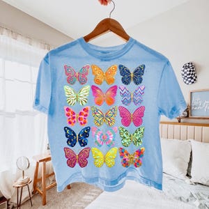 May include: A light blue t-shirt featuring a colorful butterfly design. The butterflies are in various colors and patterns, including pink, yellow, orange, and blue. The shirt is hanging on a wooden hanger.