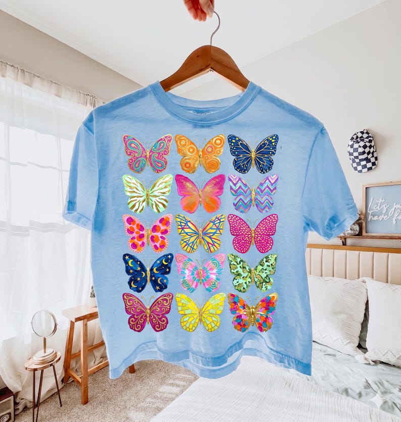 May include: A light blue t-shirt featuring a colorful butterfly design. The butterflies are in various colors and patterns, including pink, yellow, orange, and blue. The shirt is hanging on a wooden hanger.