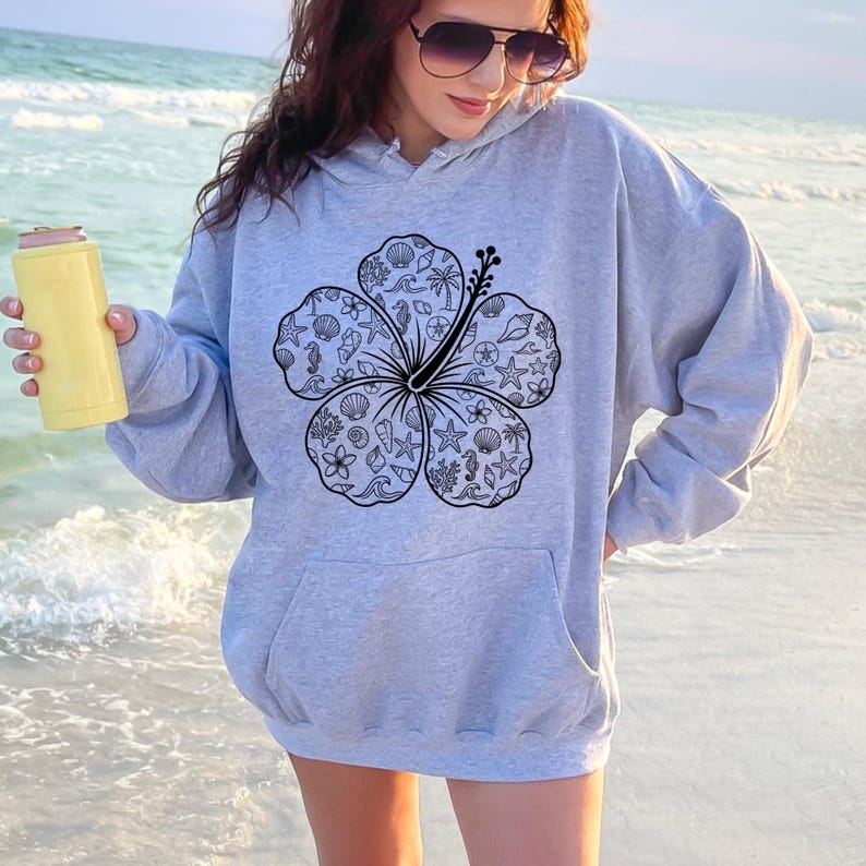 May include: A light gray hoodie with a large black and white hibiscus flower design. The flower is filled with seashell and ocean-themed illustrations. The person in the image is holding a yellow can cooler.