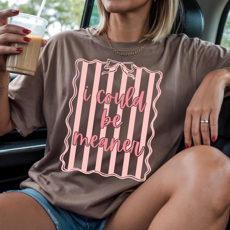 May include: A brown t-shirt with the text "I could be meaner" in pink script inside a pink and white striped design. The shirt is worn by a person.
