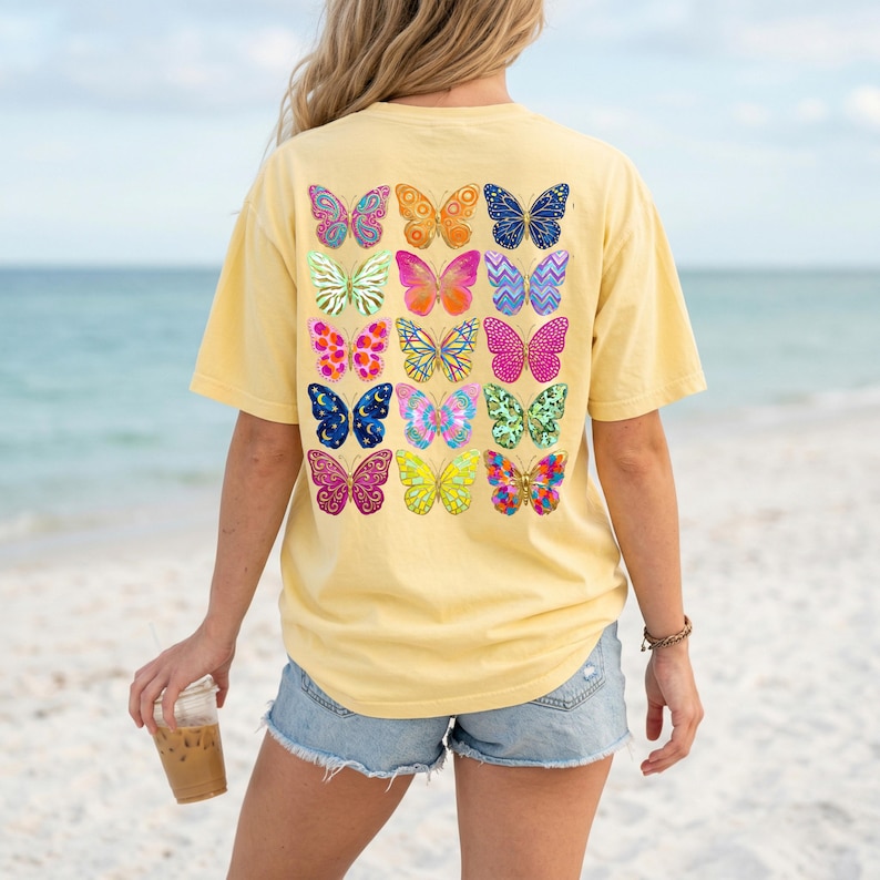 May include: A yellow t-shirt with a colorful butterfly design on the back. The butterflies are in various colors and patterns, including pink, blue, orange, and green. The shirt is a casual style, perfect for everyday wear.