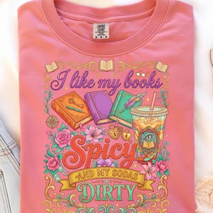 May include: Pink t-shirt with a graphic design featuring books, a drink, and the text "I like my books spicy and my sodas dirty." The design includes floral and decorative elements.