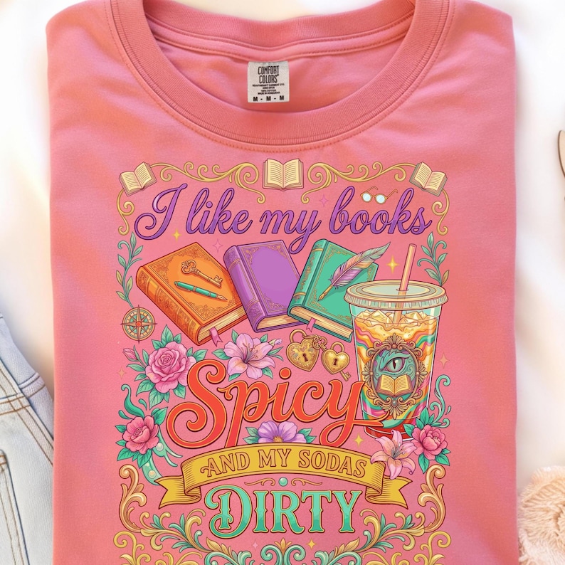 May include: Pink t-shirt with a graphic design featuring books, a drink, and the text "I like my books spicy and my sodas dirty." The design includes floral and decorative elements.