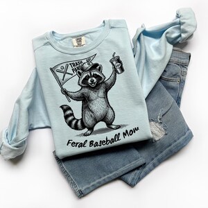 Pu&ograve; includere: Felpa azzurra con grafica di un procione che tiene una bandiera con la scritta "Trash Panda Go!" e una bibita. Il testo "Feral Baseball Mom" &egrave; sotto il procione. La felpa &egrave; abbinata a jeans di denim azzurro chiaro.