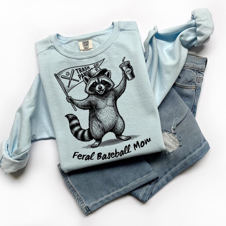 Pu&ograve; includere: Felpa azzurra con grafica di un procione che tiene una bandiera con la scritta "Trash Panda Go!" e una bibita. Il testo "Feral Baseball Mom" &egrave; sotto il procione. La felpa &egrave; abbinata a jeans di denim azzurro chiaro.