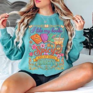 May include: Teal sweatshirt with a colorful graphic that reads "I like my books Spicy and my sodas Dirty." The design includes illustrations of books, flowers, and a drink. The sweatshirt is a comfortable, casual style.