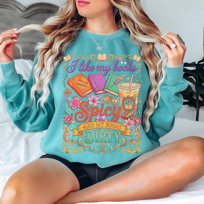 May include: Teal sweatshirt with a colorful graphic that reads "I like my books Spicy and my sodas Dirty." The design includes illustrations of books, flowers, and a drink. The sweatshirt is a comfortable, casual style.