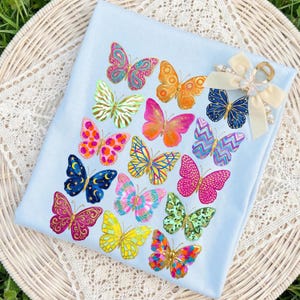 May include: A light blue fabric with a collection of colorful butterfly designs. The butterflies feature various patterns and colors, including pink, orange, gold, and blue. A decorative bow with a butterfly accent is attached to the fabric.