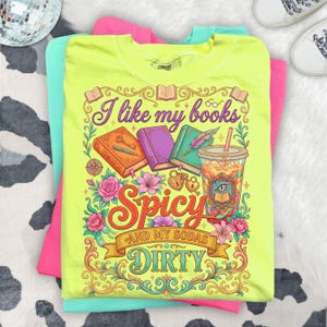 May include: A neon yellow t-shirt with the text "I like my books Spicy and my sodas Dirty" in a decorative font. The graphic includes books, flowers, and a drink. The shirt is stacked on top of a pink and a teal t-shirt.