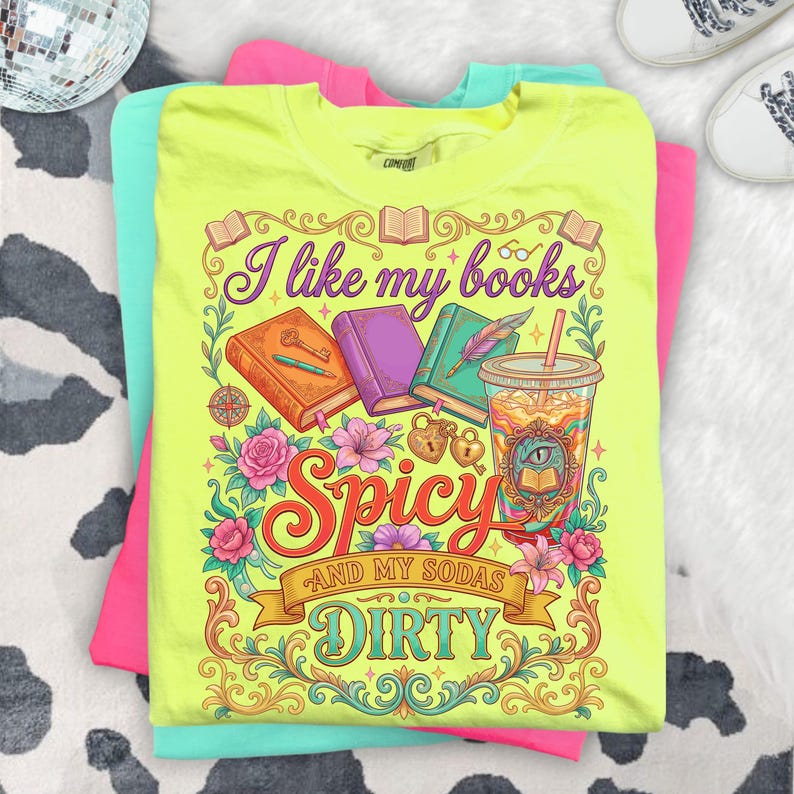 May include: A neon yellow t-shirt with the text "I like my books Spicy and my sodas Dirty" in a decorative font. The graphic includes books, flowers, and a drink. The shirt is stacked on top of a pink and a teal t-shirt.