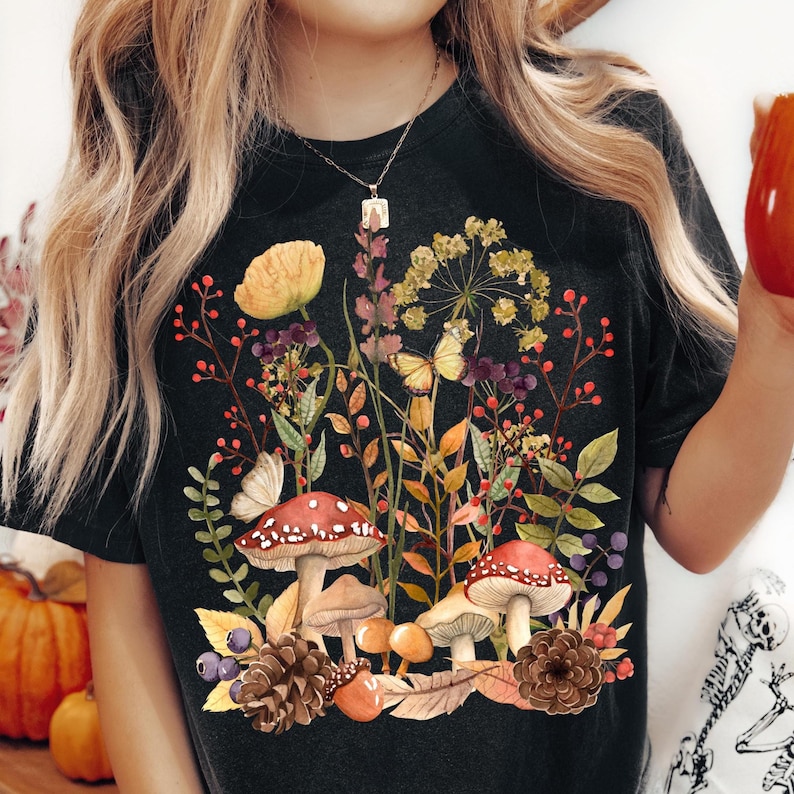 Peut inclure: T-shirt noir avec un motif aquarelle de champignons, papillons et plantes. Le motif comprend des champignons rouges &agrave; pois, des fleurs jaunes et un feuillage color&eacute;. Le t-shirt a un col rond classique.