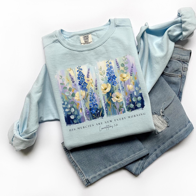 Puede incluir: Una sudadera azul claro con un dise&ntilde;o floral con flores azules, amarillas y moradas. El texto "HIS MERCIES ARE NEW EVERY MORNING Lamentations 3:23" est&aacute; impreso debajo. La sudadera se combina con unos vaqueros azules.
