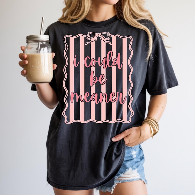 May include: Black t-shirt with the text "I could be meaner" in pink script inside a pink and white striped frame. The shirt is worn with denim shorts and gold bracelets. A glass jar with a straw and a beige drink is held in the left hand.