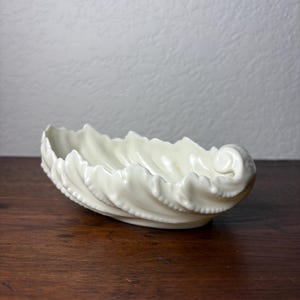 May include: An off-white ceramic bowl shaped like a curled leaf with a glossy finish. The edges of the bowl feature a scalloped, ruffled design that mimics the natural texture of foliage, culminating in a distinct spiral curl at one end.