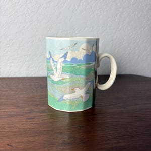 May include: A hexagonal ceramic mug featuring a decorative illustration of white seagulls flying over a stylized green and blue ocean landscape with clouds. The mug has a smooth, cream-colored handle and a multi-faceted body design.