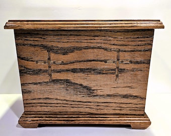Könnte beinhalten: Eine dunkelbraune Holzkiste mit einem erhöhten Sockel und zwei gravierten Kreuzen auf der Vorderseite.