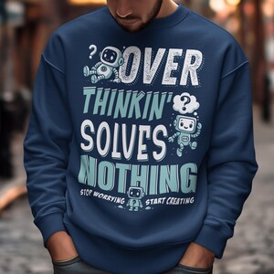 Puede incluir: Sudadera azul marino con el texto "OVER THINKIN' SOLVES NOTHING" en blanco y turquesa. El diseño incluye robots de dibujos animados y las frases "STOP WORRYING" y "START CREATING".