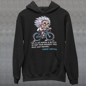 Puede incluir: Sudadera con capucha negra con una ilustración de dibujos animados de Albert Einstein montando en bicicleta. El texto dice "La vida es como andar en bicicleta. Para mantener el equilibrio, debes seguir moviéndote." -Albert Einstein