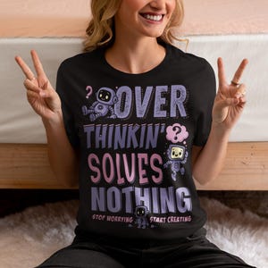 Puede incluir: Camiseta negra con el texto "OVER THINKIN' SOLVES NOTHING" en morado y rosa. La camiseta presenta gráficos de robots de dibujos animados y las frases "STOP WORRYING" y "START CREATING".